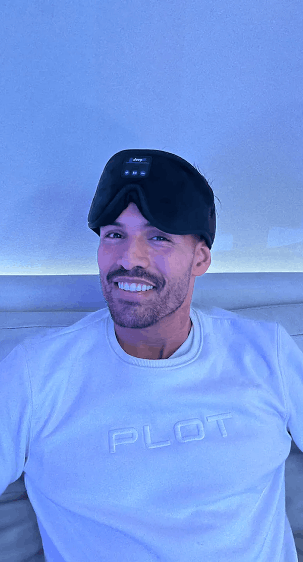 A man wearing a light gray t-shirt with 'PLOT' printed on the chest and Dreamy Sounds Sleep Mask | Bluetooth Sleep Mask, photographed in a dimly lit room with a blue cast.