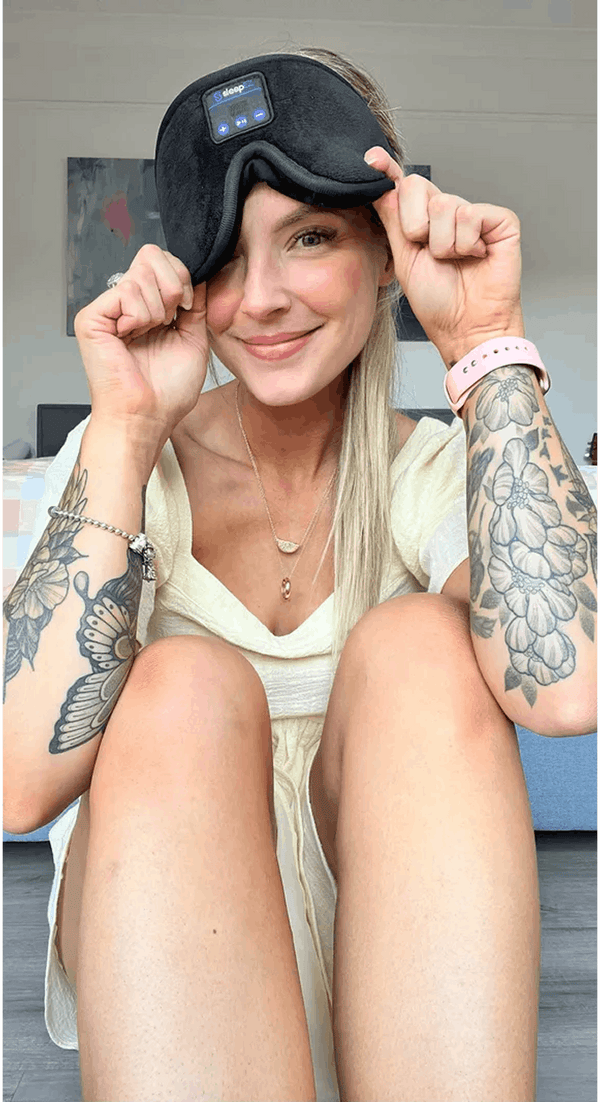 A woman with tattoos wearing Dreamy Sounds Sleep Mask | Bluetooth Sleep Mask with a digital control panel, sitting indoors.