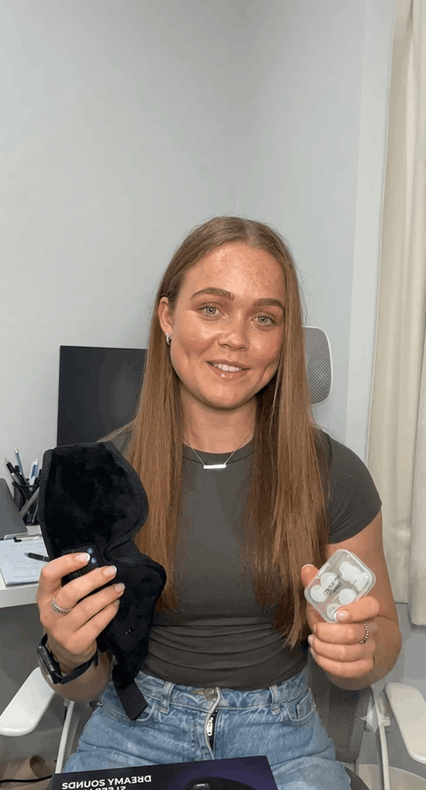 A woman holding Dreamy Sounds Sleep Mask | Bluetooth Sleep Mask and earbuds, seated in an indoor office-like setting.