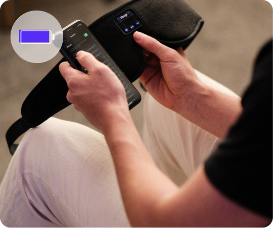 Person holding a smartphone with a battery icon overlay, trying to set up Dream Bluetooth Sleep Mask by SleepEZ