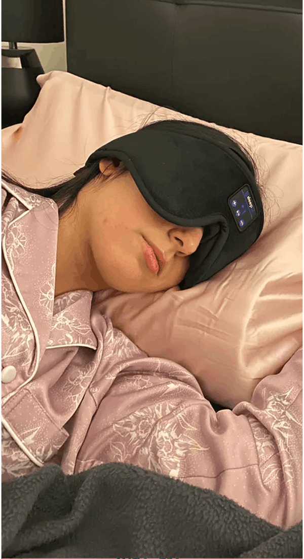 A person lying in bed wearing Dreamy Sounds Sleep Mask | Bluetooth Sleep Mask, in pink pajamas on a bed with pink satin pillows.