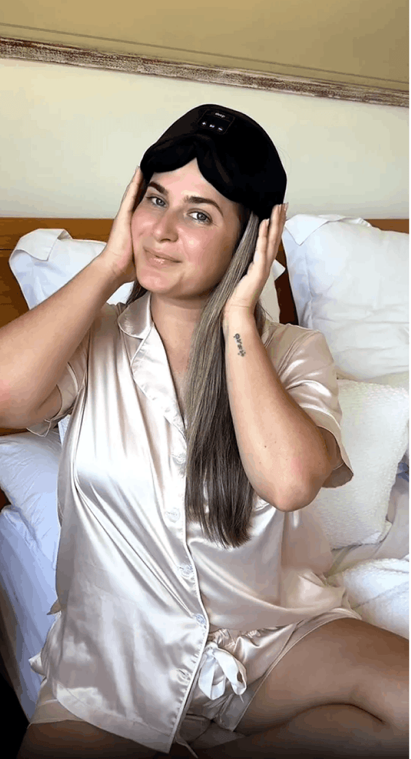A woman wearing Dreamy Sounds Sleep Mask | Bluetooth Sleep Mask and a light beige satin pajama shirt and shorts, sitting on a bed with white pillows.